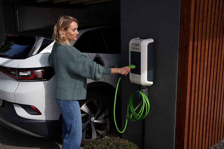 Frau bedient Wallbox an Hauswand neben einem E-Auto.