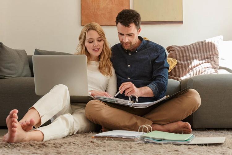 Ein junges Paar sitzt mit Aktenordner und Laptop auf dem Boden vor der Couch