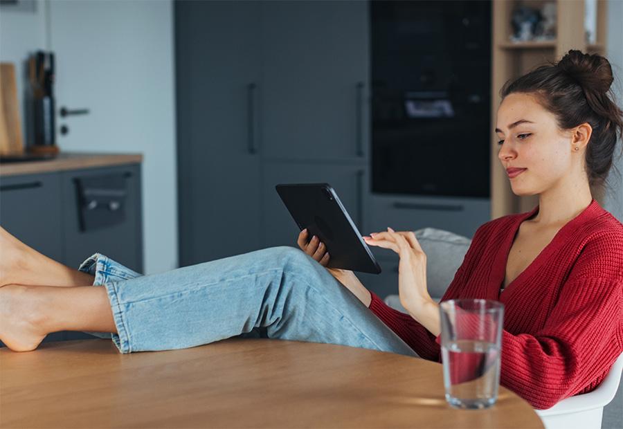 Eine Frau sitzt entspannt zuhause an ihrem Küchentisch und hält ein Tablet in ihrer Hand.