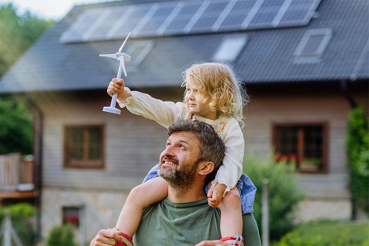 Ein Vater trägt seine Tochter auf den Schultern. Sie stehen vor einem Haus und sie hält ein Windrad Modell in der Hand.