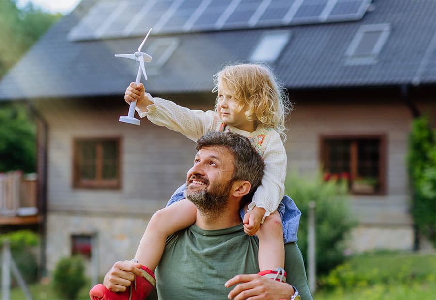 Ein Vater trägt seine Tochter auf den Schultern. Sie stehen vor einem Haus und sie hält ein Windrad Modell in der Hand.