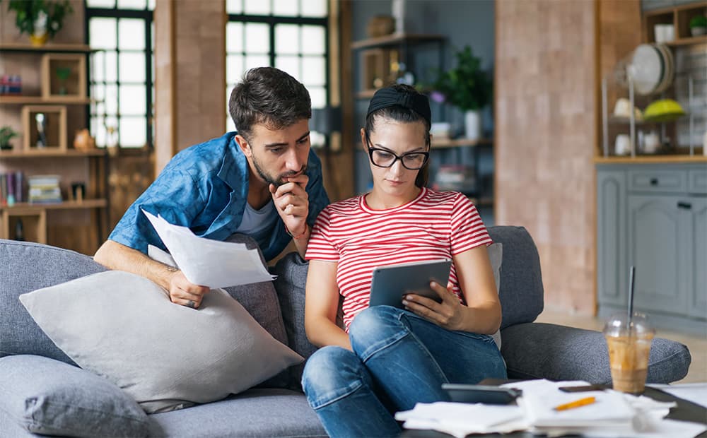 Mann und Frau sitzen im Wohnzimmer und gucken gemeinsam auf Dokumente und ein Tablet.